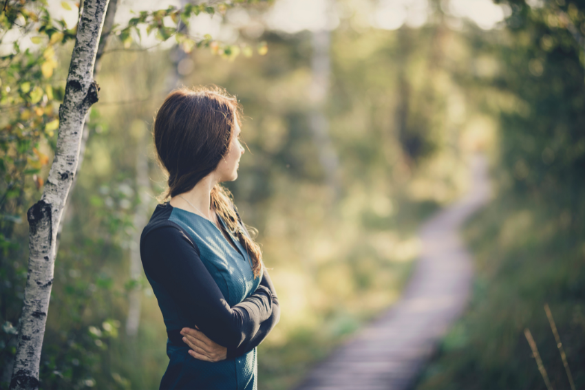 Mujer con los brazos cruzados, observando un sendero en medio del bosque con una expresión reflexiva.