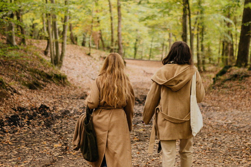 Dos personas con abrigos beige caminan juntas por un sendero en el bosque, rodeadas de árboles y hojas secas.