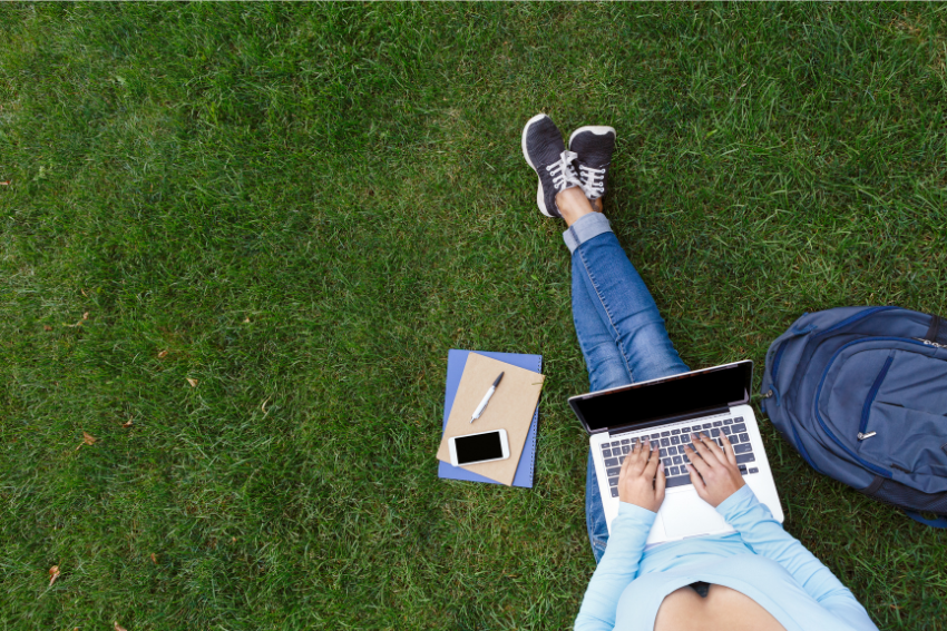 Persona sentada sobre el césped usando una laptop, con cuadernos, un teléfono y una mochila a su lado.