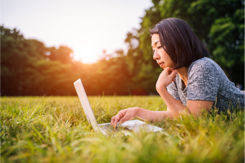 Mujer acostada en el césped escribiendo en su laptop, con la luz del atardecer iluminando su rostro.