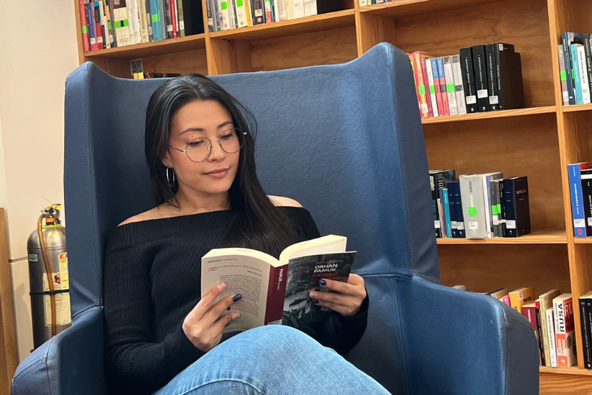 Persona con gafas y suéter negro leyendo un libro en una biblioteca, rodeada de estanterías con libros.