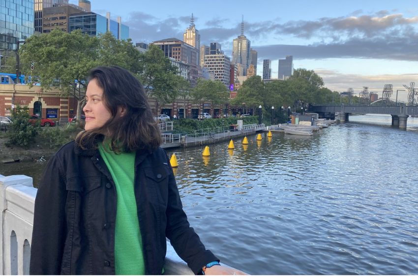 Persona con chaqueta negra y suéter verde sobre un puente con vista al río y una ciudad moderna al fondo.