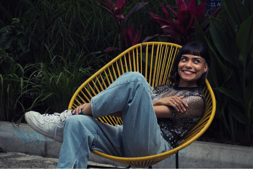 Ana Paulina, terapeuta aliada de El Diván Rojo, sentada en una silla amarilla de diseño moderno, rodeada de plantas tropicales, luciendo una blusa de lentejuelas y jeans, mientras sonríe con naturalidad.